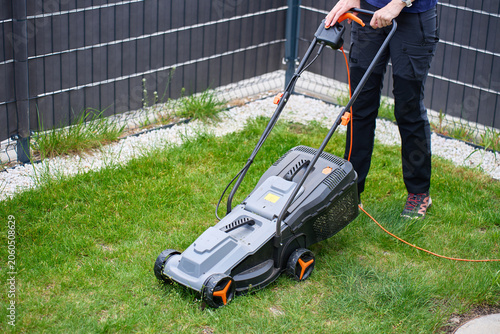 Woman operates corded lawn mower on backyard grass near fence and gravel border