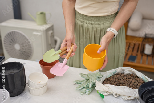 Woman prepares plant pots for indoor gardening, holding small tools and a yellow pot