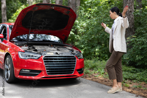 Bild auf Leinwand Woman standing next to a red car with an open hood, expressing frustration on a forest road