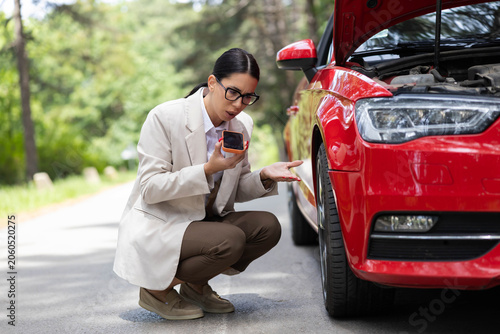 Quadro em tela Woman holding smartphone experiencing frustration with a red broken car on a roa