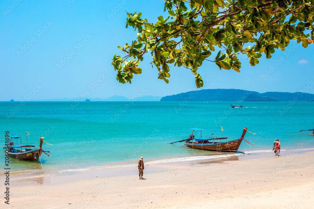 Fototapeta premium View of a sandy beach with longtail boats and turquoise water through tree branches. tropical landscape, summer vacation, Krabi marine tourism.Krabi, Thailand, March 20, 2024