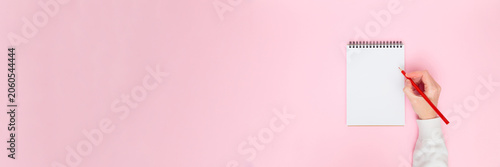 Female hand writing on blank notebook on pastel pink background. Minimal flat lay banner with copy space for notes, planning, education, business, blogging, creative content