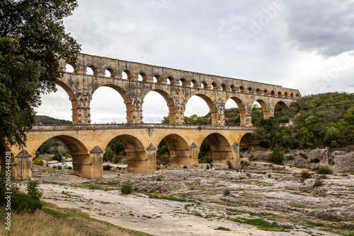 The magnificent Pont du Gard Roman aqueduct spans a dry riverbed in southern France under a dramatic, cloudy sky, framed by trees