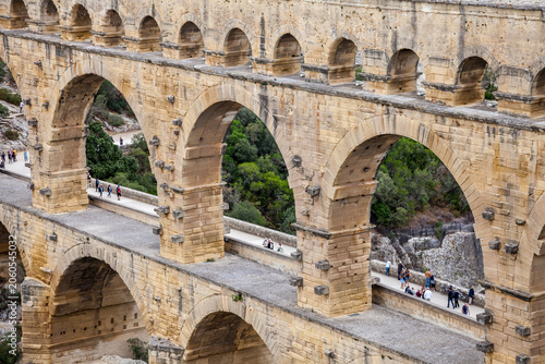 Tourists walk across the ancient three-tiered Roman aqueduct Pont du Gard in southern France, with golden limestone arches framing green valley.