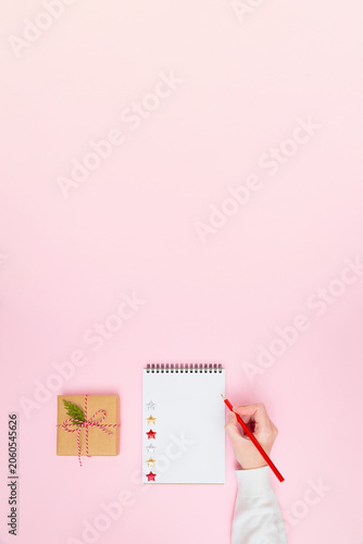 Female hand writing on blank notepad with gift box on pastel pink background, festive flat lay vertical banner with copy space for holiday planning, goals, to-do list or wish list