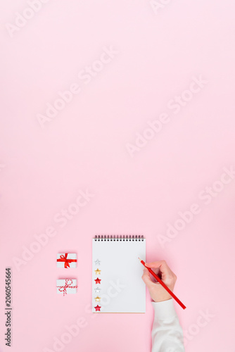 Holiday vertical banner with female hand writing in blank notebook, Christmas gifts and glitter stars on pink copy space for planners, checklists, marketing designs and festive social media content