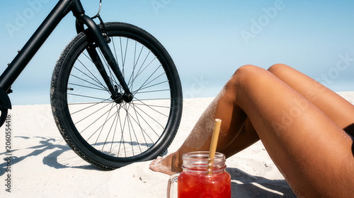 Tanned legs next to a black bicycle and red drink in mason jar on sandy beach