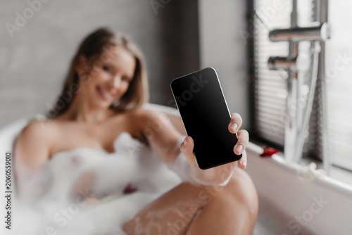 Konstfotografi Smiling Young Woman Enjoying Relaxing Bath While Holding Smartphone With Empty Screen, Seated In Bathtub, Bubbles Present, Home Setting