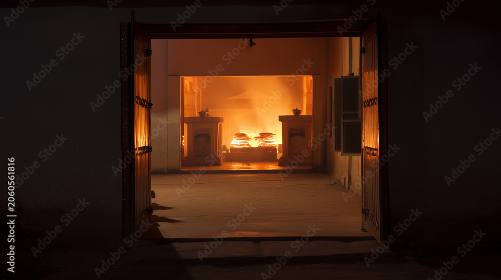 Fototapeta premium Respectful Hindu cremation ceremony scene at night, viewed from a distance through an open architectural doorway, warm orange ceremonial fire glowing inside a simple crematorium or