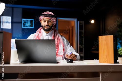 Middle Eastern man wearing Arabian clothing is seated at the table, engrossed with research on his digital laptop.