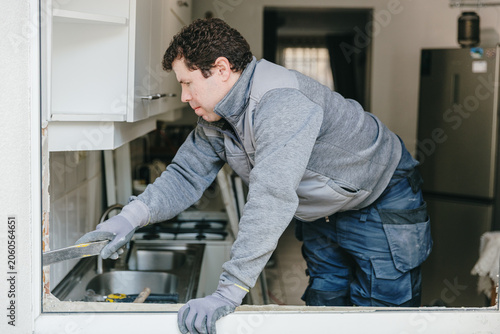 Foto A portrait of a mature handyman in grey gloves using a crowbar to loosen a corner of an old plastic window frame before dismantling it