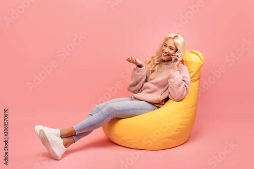 Tableau sur toile Smiling Young Woman Engaged In Conversation On Smartphone While Sitting Comfortably On Yellow Bean Bag In Modern Studio Setting With Pink Background