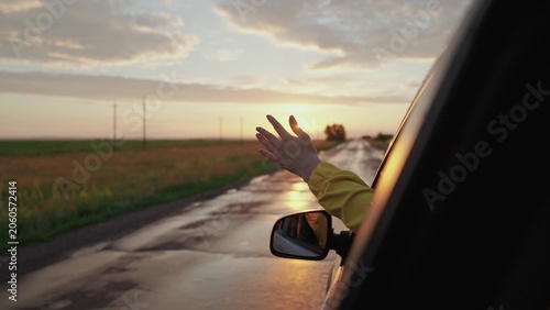 Canvas Print Girl in car window waves her hand and catches wind at sunset, Wet road after rain