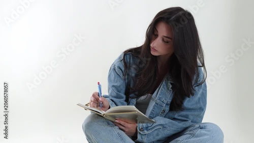 Creative writing and education concept with a young woman student diligently writing in a notebook with a pen. The model is casually dressed in denim, sitting on the floor, and isolated on a clean whi
