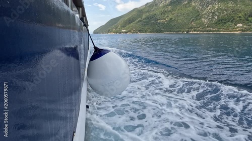 Close-up view from the side of a ferry cruising through calm blue waters near Lefkada, Greece. Sea foam trails along the hull while green mountains and coastline pass by in the background on a sunny d