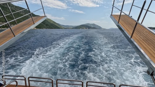 Symmetrical rear view from a passenger ferry sailing through the Ionian Sea in Greece. White foamy wake stretches across deep blue water toward distant mountainous islands, framed by the ferry loading