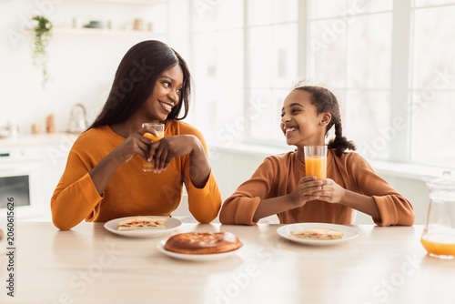 Foto Happy Black Female Adult And Young Girl Smiling While Enjoying Breakfast Together At Kitchen Table, Drinking Juice And Eating Food In Modern Home Setting