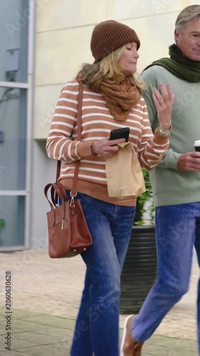 Vertical video: Entering frame couple walking to bench at glass bldg, with phone, paper bag and cup