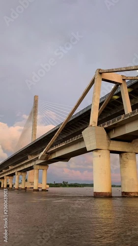 Low-angle view of a concrete Pumarejo bridge crossing a wide Magdalena river at sunset, with calm water, massive support pillars, and pastel clouds creating a moody architectural landscape.