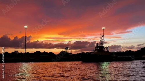 Vibrant orange and purple sunset sky over calm water, with dark silhouetted shoreline, tall light pole, and reflections creating a dramatic evening seascape.