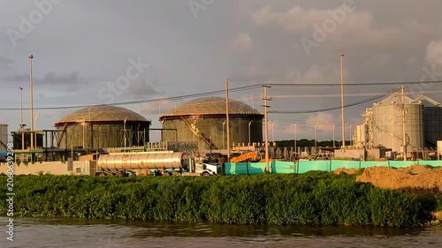 Industrial facility with large storage tanks and processing structures near a riverbank, showing wastewater treatment infrastructure, pipelines, and heavy industry under a cloudy sky.