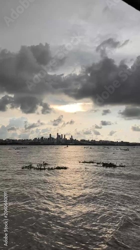 Moody sunset scene with dark clouds, glowing sky, and distant city skyline reflected on calm water, creating a dramatic urban seascape atmosphere.