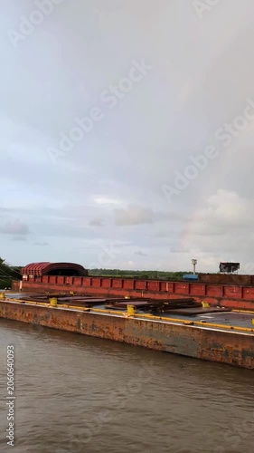 Massive red cargo vessel moored in a muddy river channel near the shore, with towing cables visible under an overcast sky. Industrial maritime transport scene.