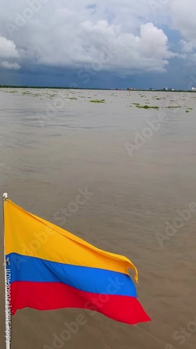 Colombian flag waving on the stern of a moving boat, with rippling river water and wake in the background. Travel and national identity concept with scenic maritime motion.