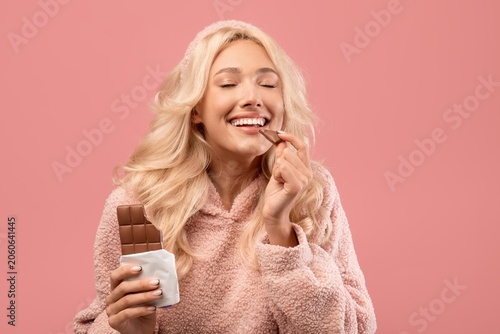 Fotografía Smiling Young Woman Holding Chocolate Bar And Enjoying A Piece, Happy Expression On Face, In Studio Setting With Pink Background