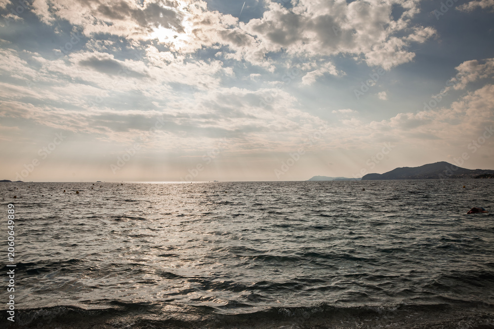 Fototapeta premium Panoramic view of the Mediterranean Sea off Hyeres on the French Riviera, in france, sunlit clouds, distant coastline, small buoys, hazy horizon and textured waves along the shore in southern France.