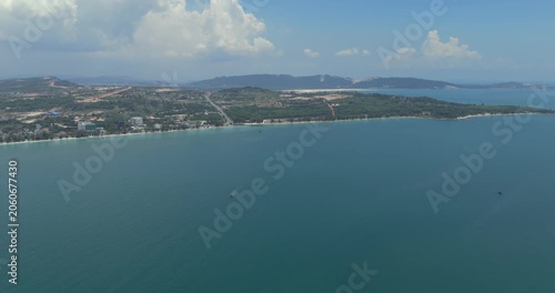 Forward aerial view of a calm, vast blue sea with boats cruising near a green, hilly coastline under a cloudy sky.