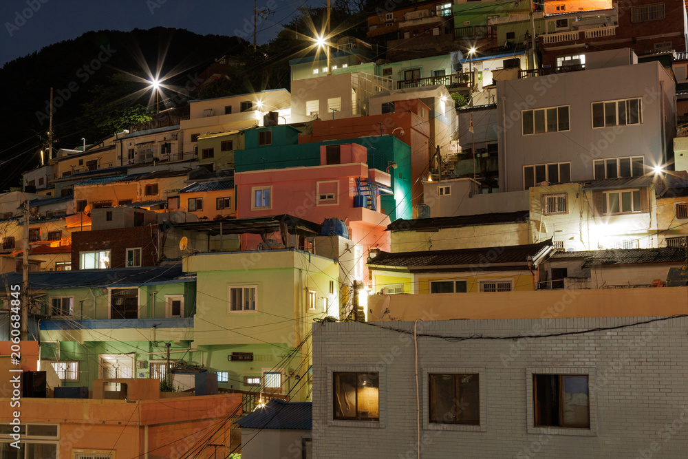 Fototapeta premium Brightly illuminated colorful houses on a hillside at night in Busan, South Korea. Dense residential neighborhood with glowing windows, city lights and mountain backdrop.