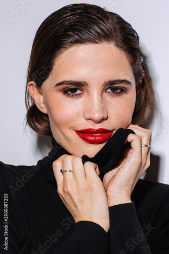 A confident young woman with brunette hair and bold red lipstick poses in a black turtleneck sweater, gently holding the collar near her face. Studio lighting highlights her smooth skin, beauty look
