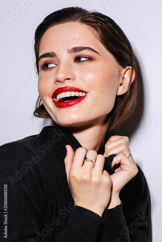 Joyful young woman in a beauty portrait with red lipstick, brunette wet hair, bright smile and black turtleneck, posing with rings under bold studio lighting against a white background.
