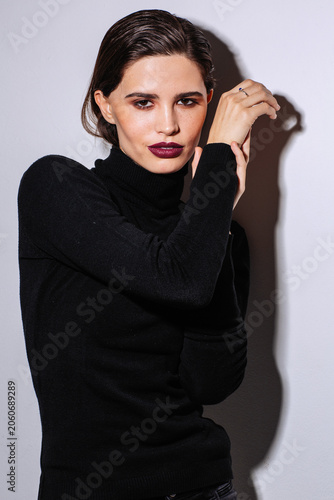 A stylish young woman in a black turtleneck poses with elegant hands, bold plum lipstick, polished makeup, and a confident expression against a minimalist studio background. Dramatic side lighting 