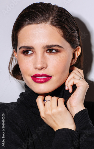 A young woman with flawless skin and vivid pink lipstick poses in a black turtleneck sweater against a light studio background. She gently holds the collar while looking aside