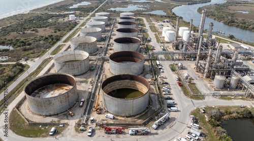 An aerial view of a strategic petroleum reserve facility with rows of large oil storage tanks appearing partially depleted, surrounded by maintenance vehicles and refinery infrastructure
