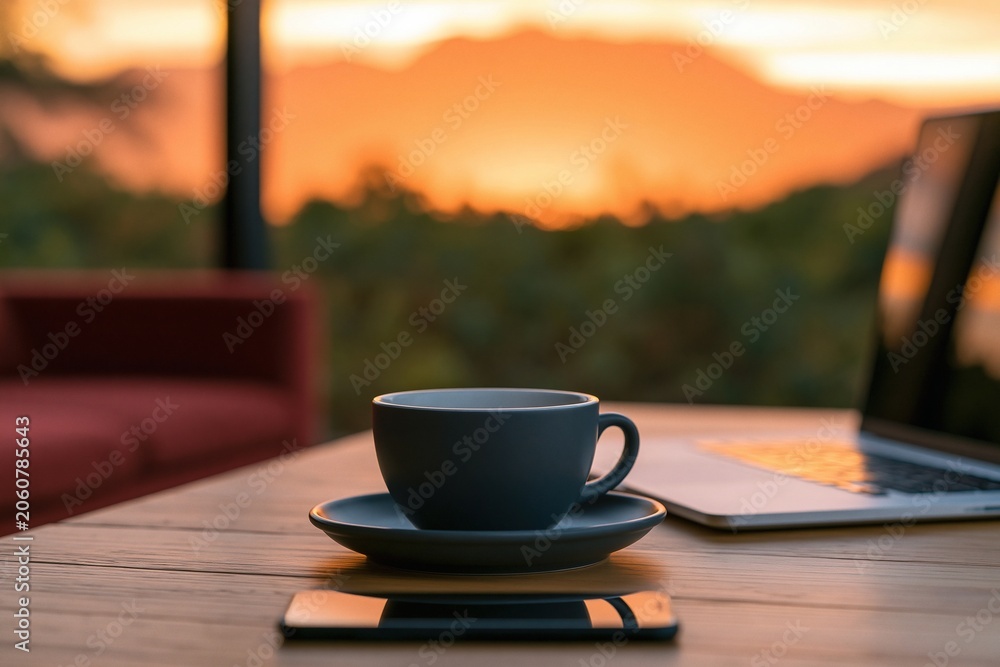Fototapeta premium Coffee cup and laptop on a wooden desk with a smartphone at a beautiful sunset