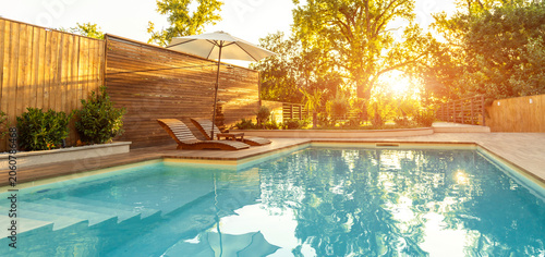 Panorama of luxury swimming pool in Modern Villa with Palm Trees and wooden lounge chairs at sunset