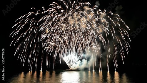 A nighttime scene featuring vibrant fireworks exploding over water, reflecting warm golden light