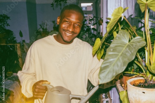 Smiling young man watering lush green houseplants in a sunlit room, nurturing indoor garden plants for a peaceful wellness and domestic lifestyle