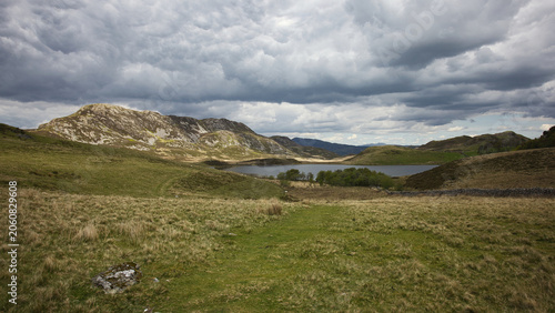 A scenic view of Cregennen lake in North Wales nestled among rolling green hills and rugged rocky mountains under a dramatic, cloudy sky.