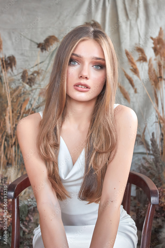 Fototapeta premium A young woman with long, wavy brown hair and expressive blue eyes. She is wearing a white sleeveless top and is looking directly at the camera.