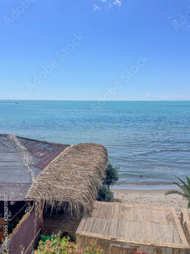 Vertical high angle photo of a rustic beach bar bungalow with a dry straw thatched roof overlooking a calm turquoise sea on a bright sunny day