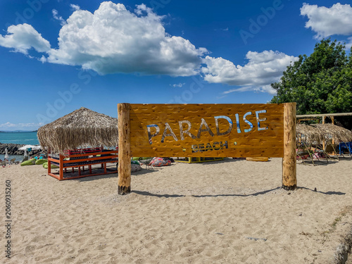 Horizontal photo of a large wooden sign with Paradise Beach text on a sandy shore with tropical thatched bungalows under a blue sky with fluffy white clouds