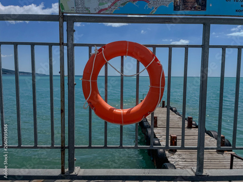 Bright orange lifebuoy hanging on a metal fence with a wooden pier and calm turquoise sea in the background on a sunny day