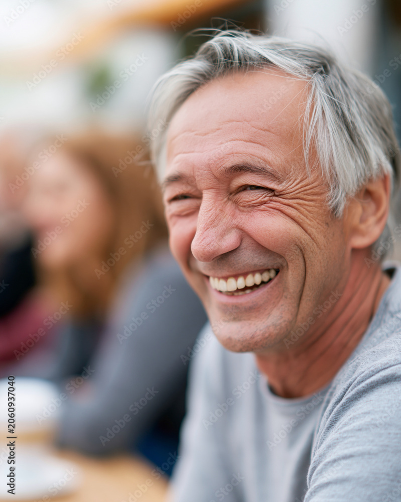 Gray-haired man smiles warmly at the camera. He sits at a cafe table with blurred background figures. His expression radiates joy and relaxed happiness. Ideal for lifestyle, retirement
