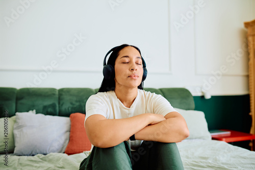 Vászonkép Young woman sitting on a bed with closed eyes, wearing headphones, finding inner