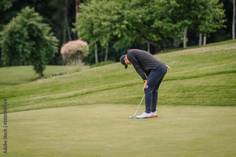 Obraz premium Male golfer in black long-sleeve shirt and navy pants prepares to putt on green golf course surrounded by trees and manicured grass in a sunny outdoor setting