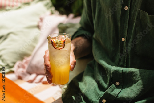 Close-up of a person in a green linen shirt holding a vibrant, layered tropical lemonade garnished with a passion fruit half while relaxing on a cozy outdoor terrace.